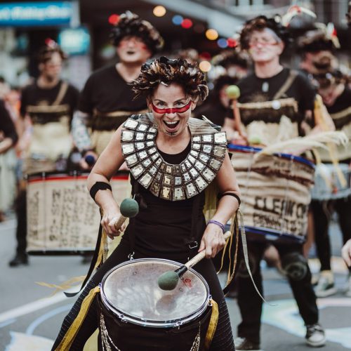 Group of batucada drummers marching down the street, the shot is focussed on one performer who's enthusiastically grinning at the camera