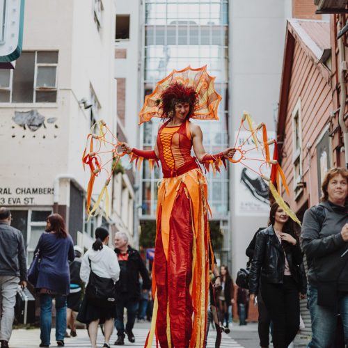Performer on stilts in a red and orange costume walking down the street