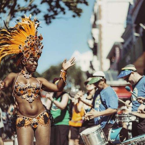 Batucada dancer performing in a sunny street, with drummers in the background