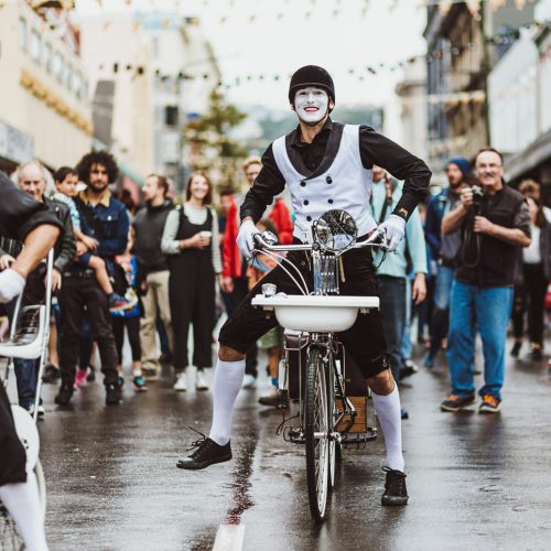 A smiling mime riding a bicycle down the street