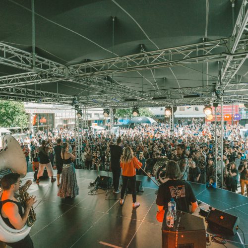 Shot looking over the shoulders of a brass band performing on stage with a large crowd watching