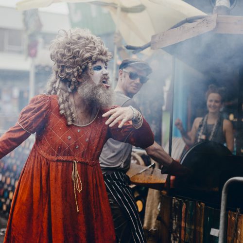Street performer with a big wig in a red dress and white face paint pulling a face
