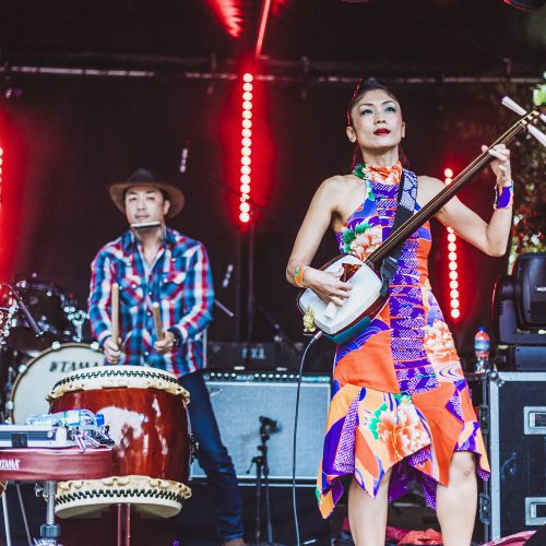 A woman in a vibrant costume playing and singing, with a drummer in the background, at the glover park stage