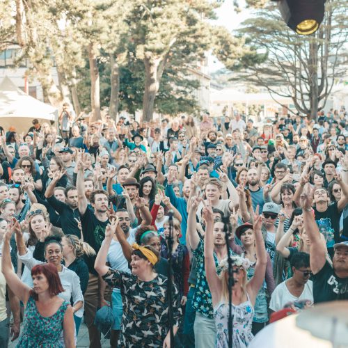 Large crowd at daytime stage with their hands in the air