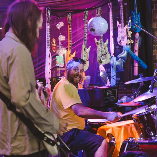 Drummer on stage smiling at the camera with band mates in the background