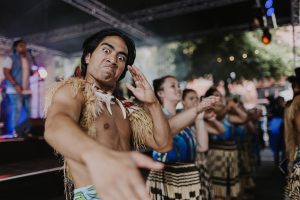 Performer looking into the camera, while men and women do a traditional Pasifika dance in the background