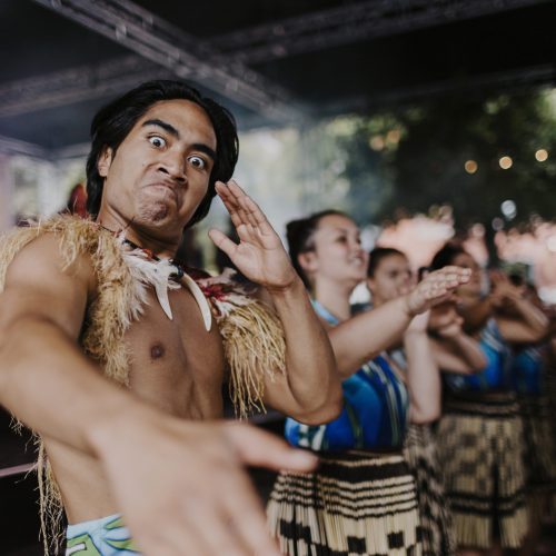 Performer looking into the camera, while men and women do a traditional Pasifika dance in the background