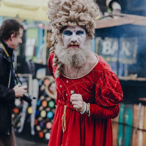 Street performer with a big wig in a red dress and white face paint looking into the camera